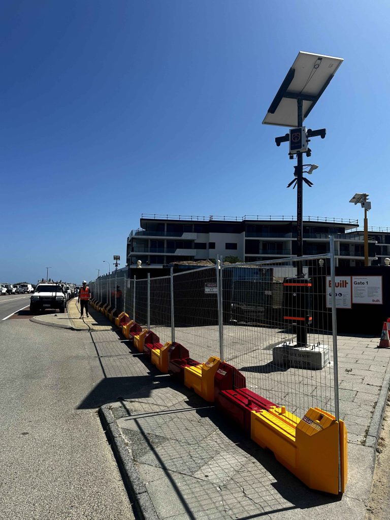 Gallery image: Construction site with fencing, red and yellow barriers, and solar-powered lights along a street. Building under construction visible.