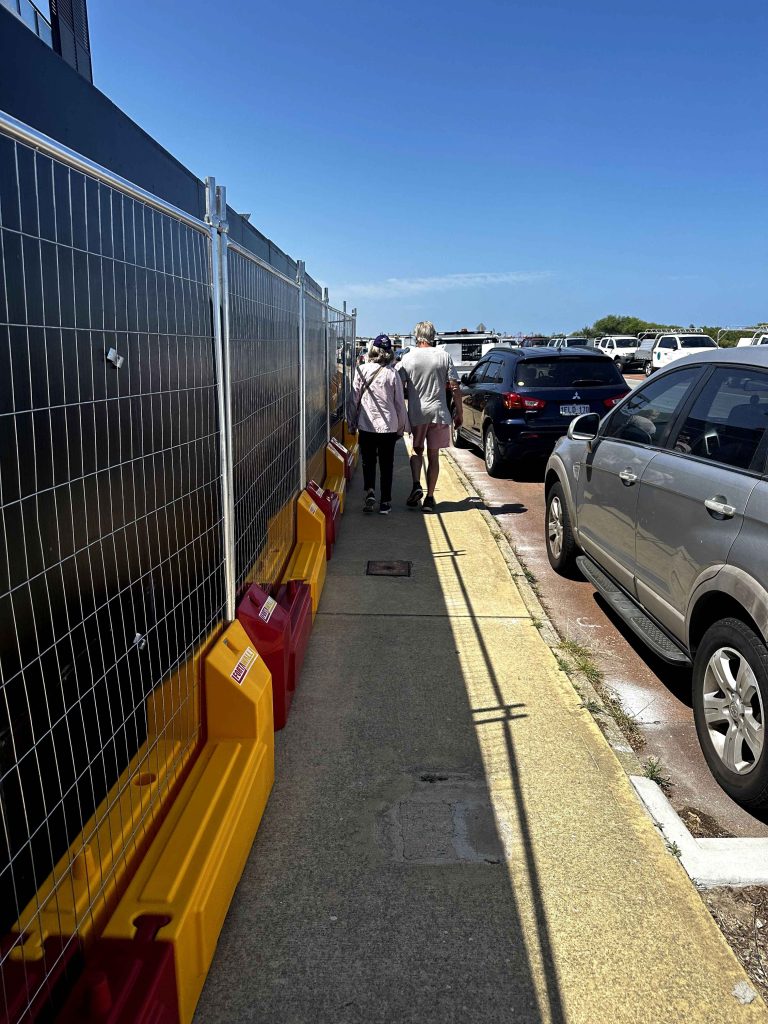 Gallery: Couple walks on a sidewalk next to a construction fence and parked cars under a clear blue sky. The scene is brightly lit.