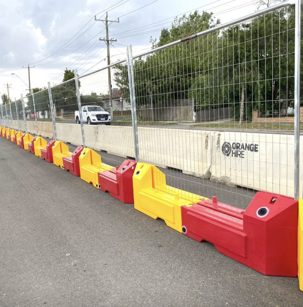 Gallery: Temporary fencing with red and yellow barriers along a road. A white truck is visible behind the fence. Orange Hire is printed on the barrier.