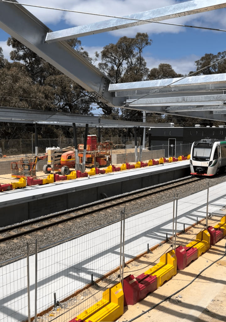 Gallery: Train at a station under construction. Yellow and red barriers line the platform. Construction equipment is present. Steel beams overhead.