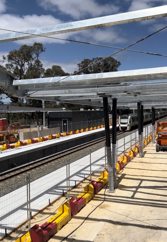 Gallery: A train approaches a station platform with yellow & red barriers. Overhead, a steel structure provides shade. Trees & blue sky visible.