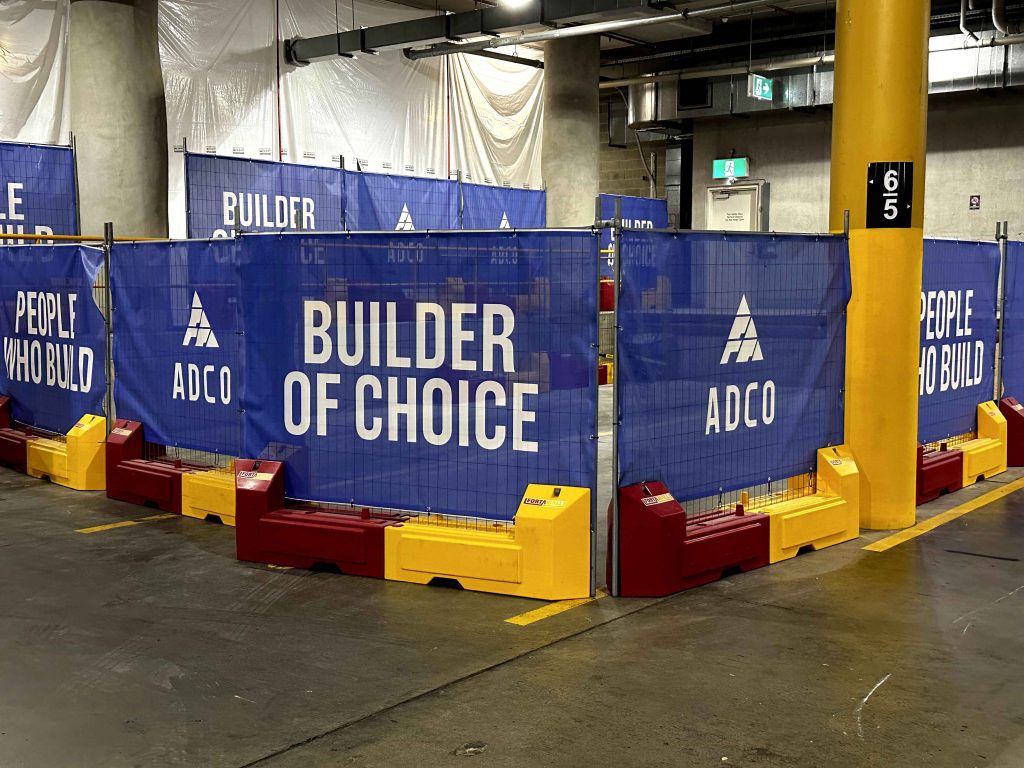 Gallery: ADCO Builder of Choice construction site barrier with blue mesh, white text, and red/yellow base. People who build visible. Column 65 is nearby.
