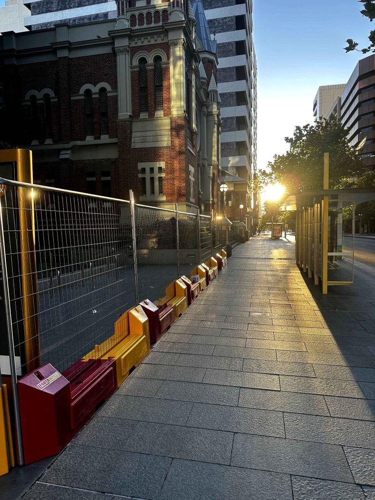 Gallery image: City street view with sunlight shining between buildings, casting shadows on the sidewalk. A fence runs along the side of the street.