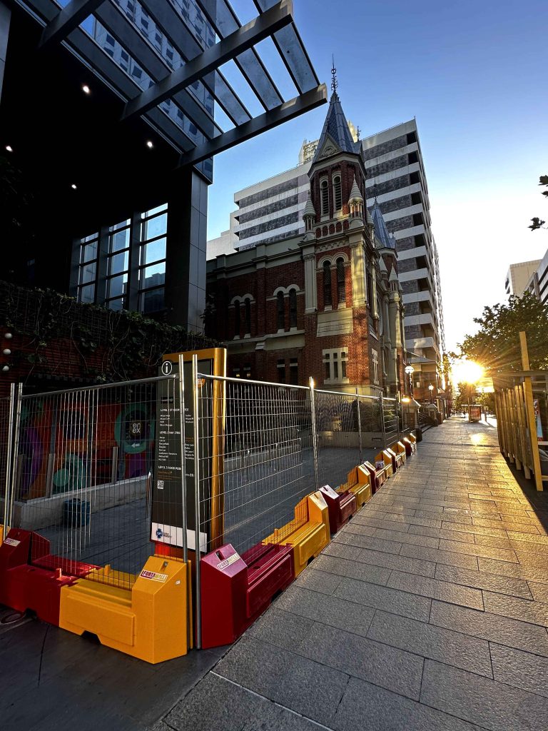 Gallery: Urban landscape with a red brick cathedral between modern buildings, a fenced construction site, and sunlight on a stone-paved street.