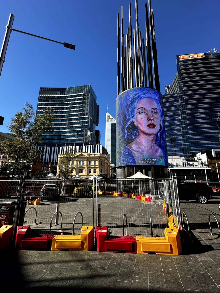 Gallery image of a public art installation featuring a portrait of Kathy Sirma on a cylindrical screen with metal rods extending upward.