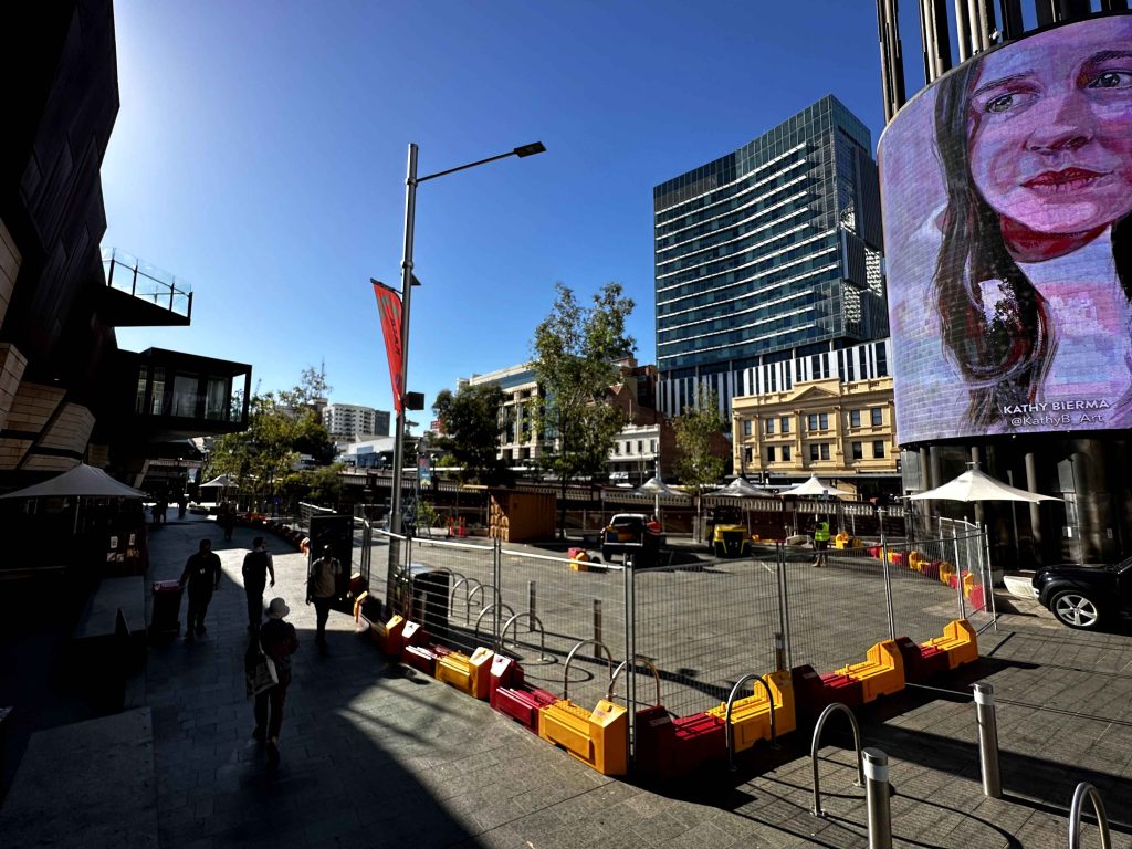 Gallery: Perth, Australia city scene. Modern architecture, a digital portrait, and construction barriers under a clear blue sky.