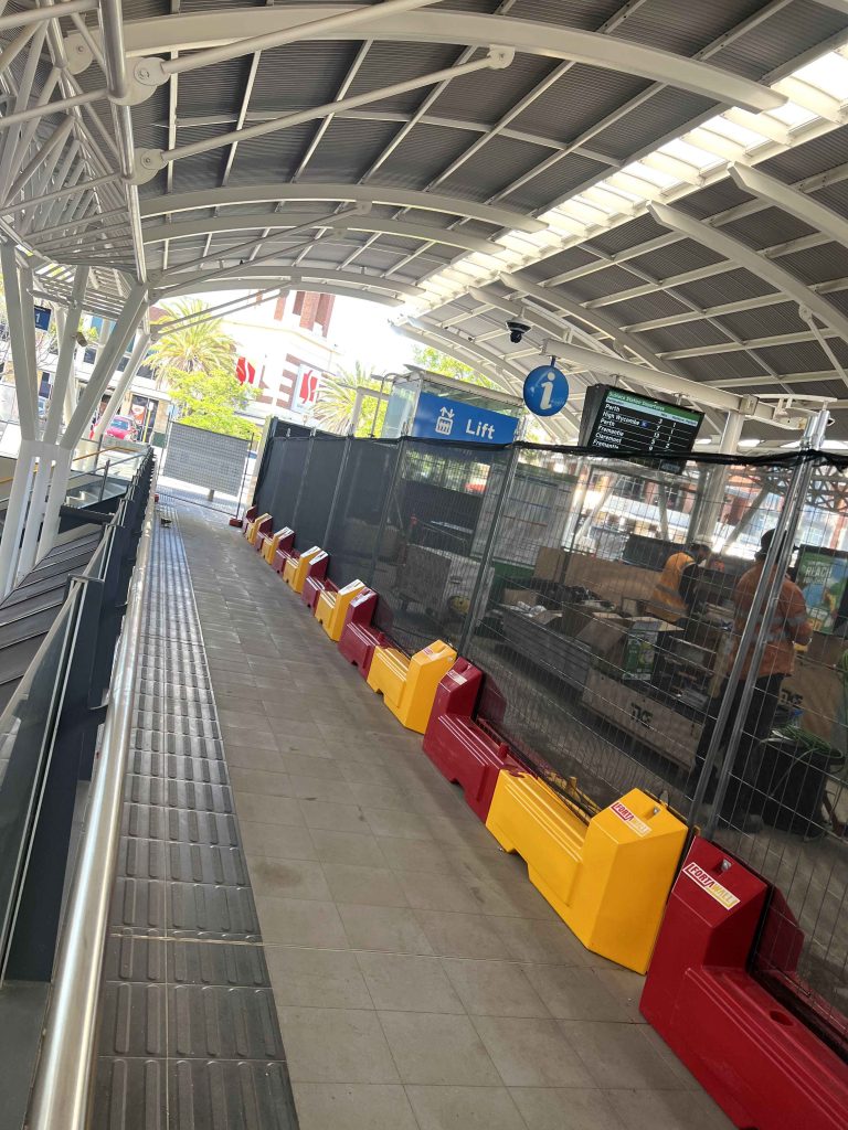 Gallery image: Construction barriers line a train platform. A lift sign is visible, along with a train schedule display. The platform is under a metal roof.