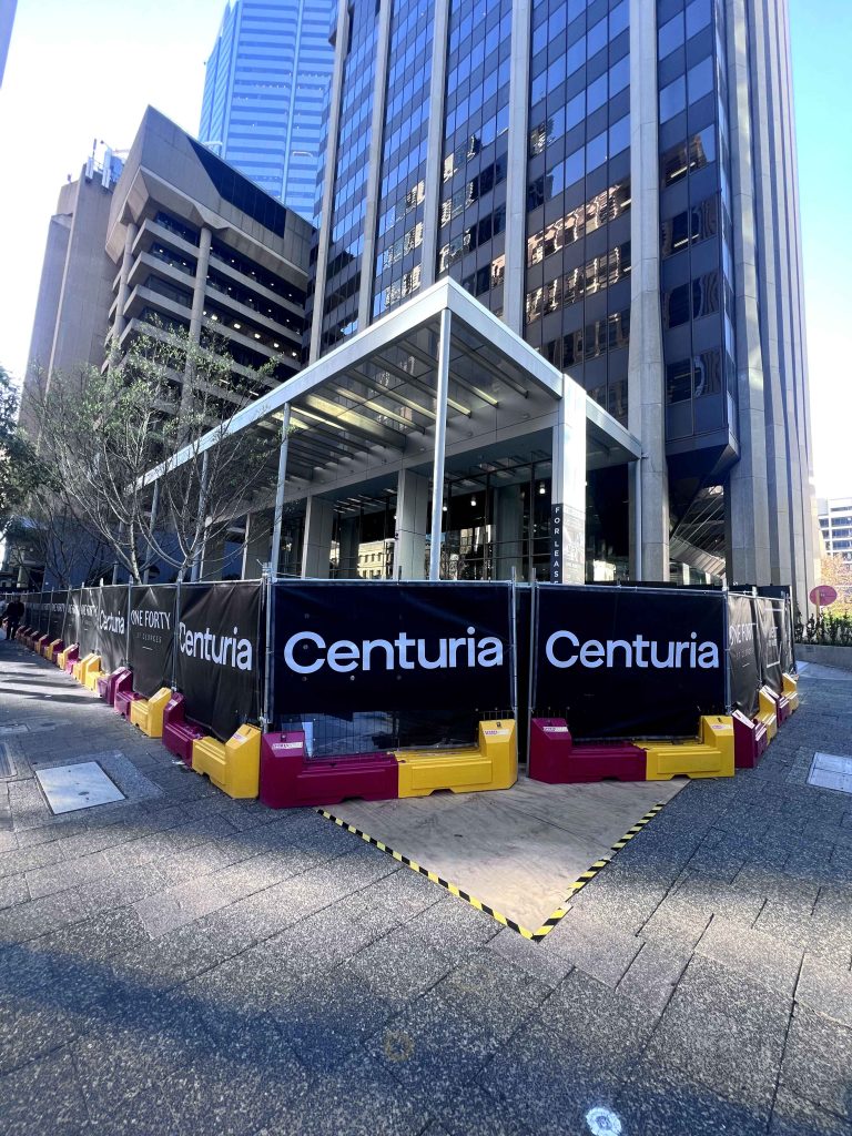 Gallery: Centuria signage around a construction site in front of a tall building with a glass facade on a sunny day.