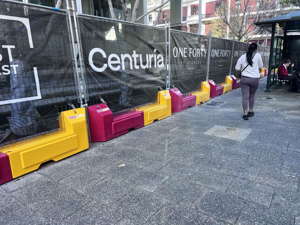 Gallery image: Construction site barrier with Centuria signage on a city sidewalk. A woman walks by, and a bus stop is visible in the background.