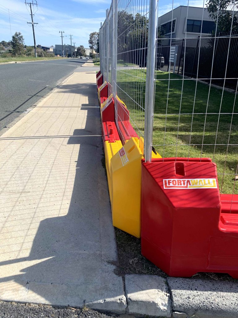 Gallery: A temporary fence with red and yellow plastic bases runs along a sidewalk. The fence is made of metal wire mesh and is used for construction.