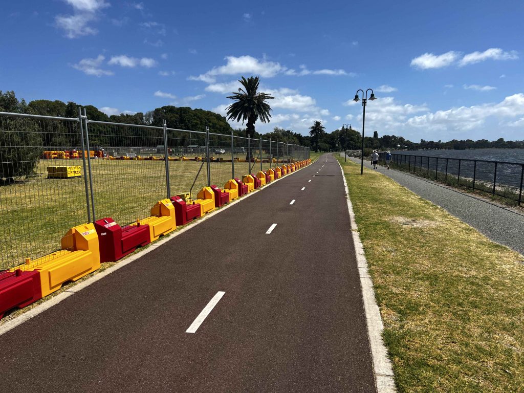 Gallery image: Path with red and yellow barriers alongside a fenced park area. A waterfront and blue sky with clouds are visible.