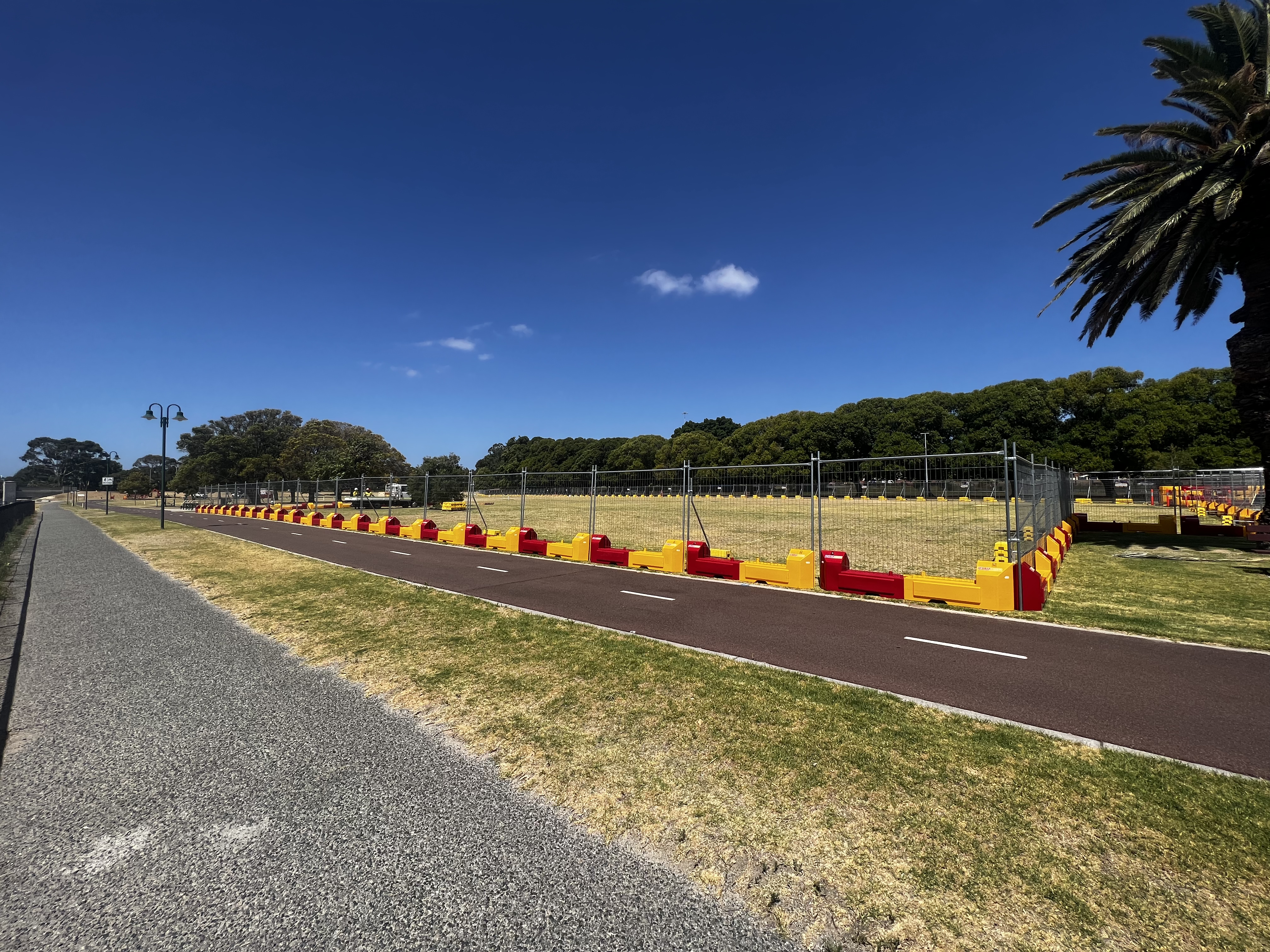 About: Fenced field with red and yellow barriers, a path, and a palm tree under a clear blue sky. Trees line the background.