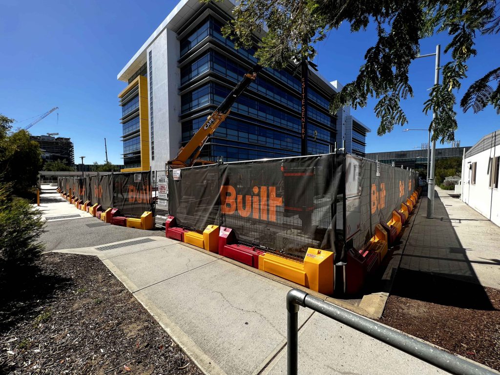 Gallery: Construction site with a modern building, crane, and black fence featuring the word Built in orange. Red and yellow barriers line the fence.