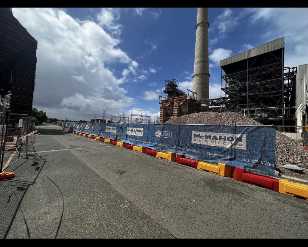 Gallery: Industrial demolition site with tall chimney, metal structures, and debris pile behind a McMahon Services safety barrier on a city street.