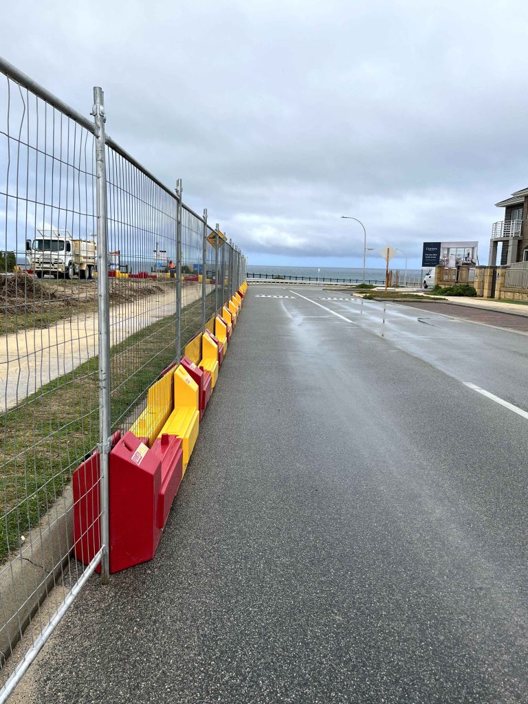 Gallery: Construction site with a metal fence and red/yellow barriers along a wet road near the ocean under a cloudy sky.