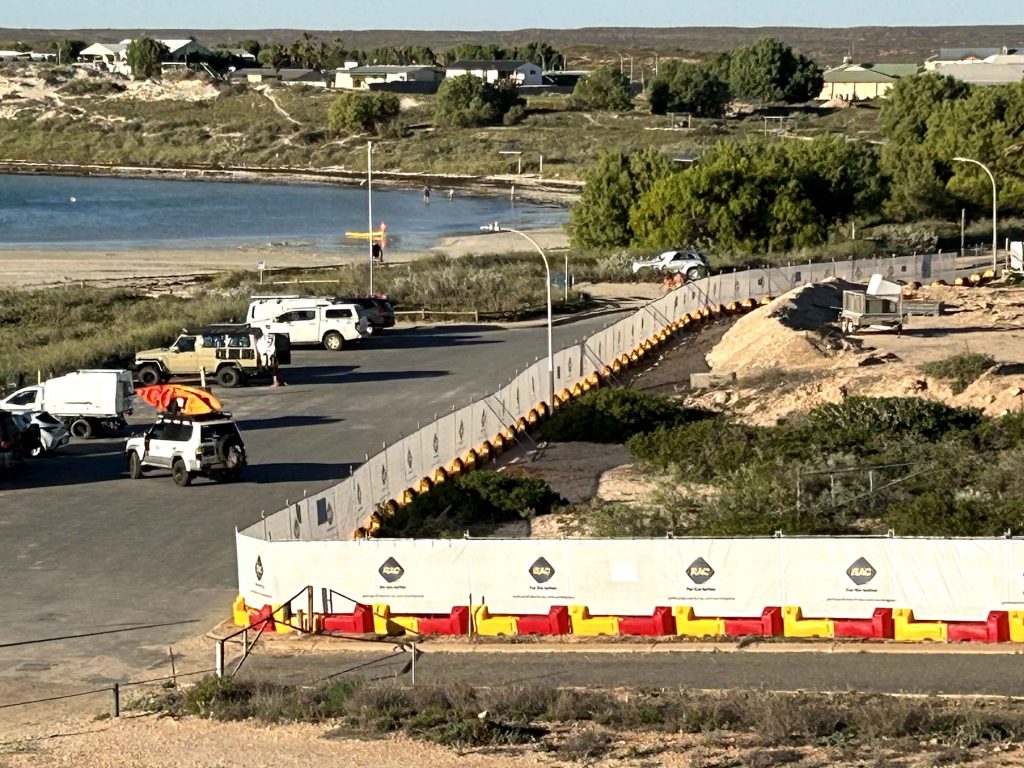 Gallery image: Coastal parking lot with 4x4 vehicles near a beach and lagoon. A construction barrier is visible, with buildings and vegetation in the background.