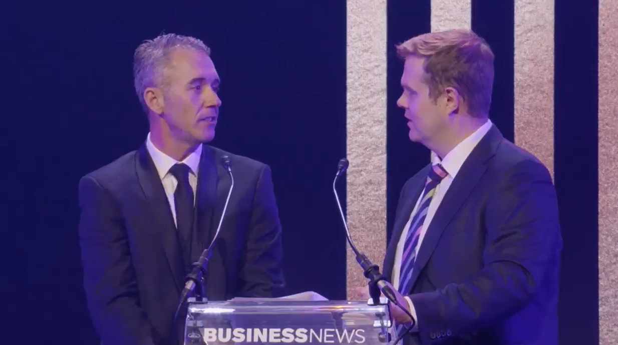 Two men in suits at a Business News event. One man is speaking into a microphone, while the other is listening attentively.